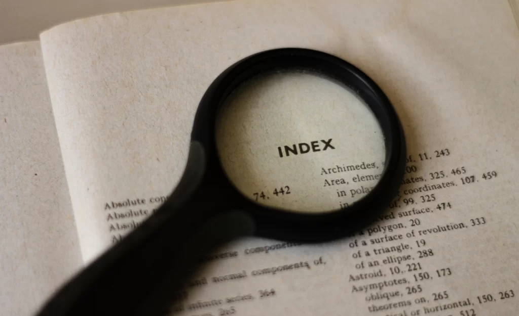 Uma lupa ampliando a palavra "INDEX" em uma página de livro, destacando uma lista de tópicos, a lupa simboliza a procura de uma palavra-chave.